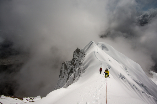 Climbers ascending a snowy mountain ridge under a cloudy sky