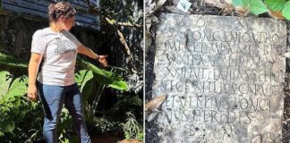 A woman in a garden pointing at a Roman inscription on a stone slab surrounded by greenery