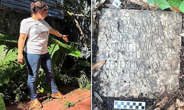 A woman in a garden pointing at a Roman inscription on a stone slab surrounded by greenery