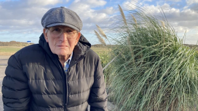 Elderly man in a black jacket and cap sitting outdoors near tall grass