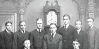 Group portrait of men associated with the Skull and Bones secret society, featuring a skull and crossed bones on a table