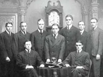 Group portrait of men associated with the Skull and Bones secret society, featuring a skull and crossed bones on a table